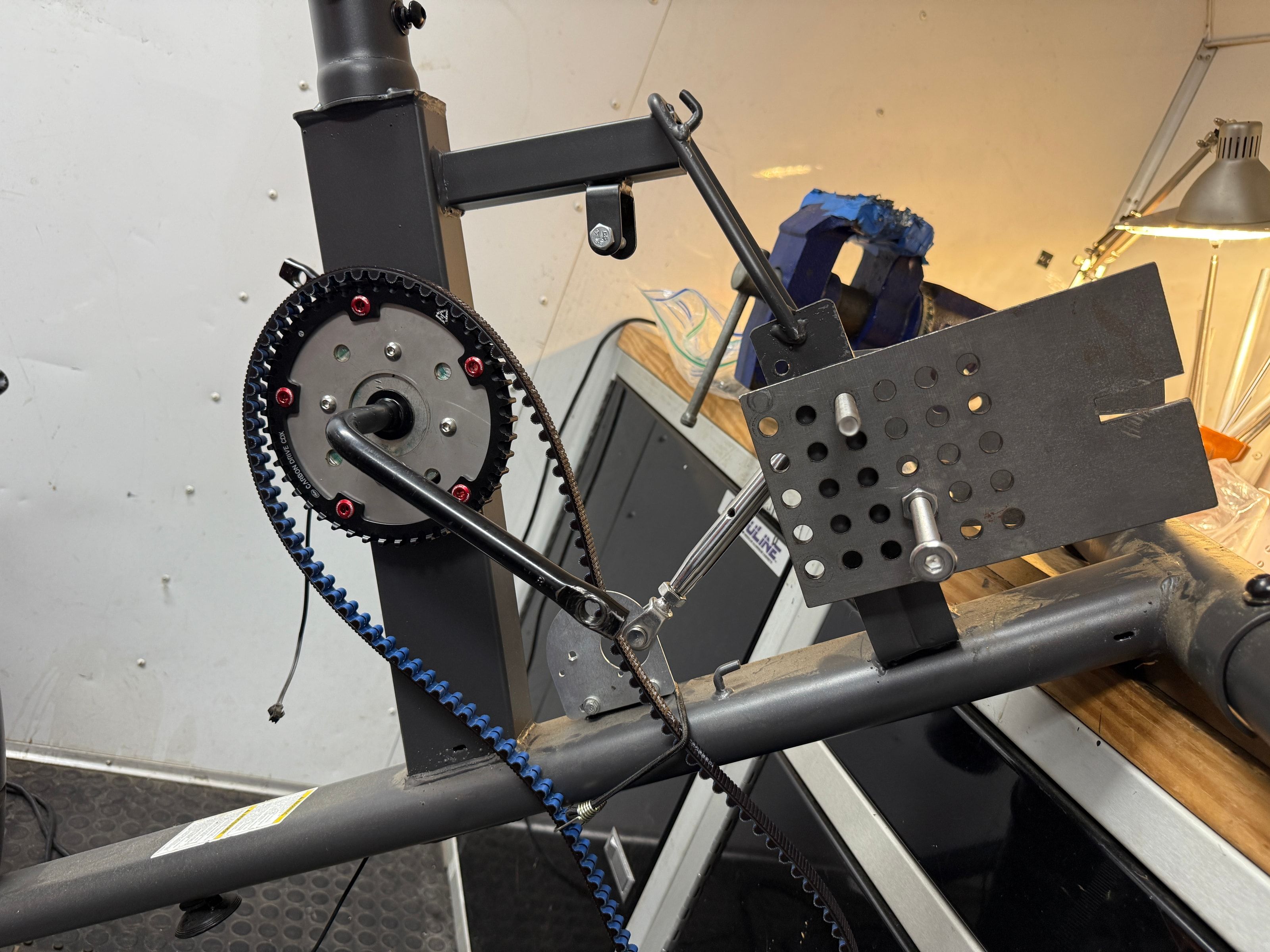 Perforated black prototype plate clamped beside a Schwinn crank and blue toothed belt